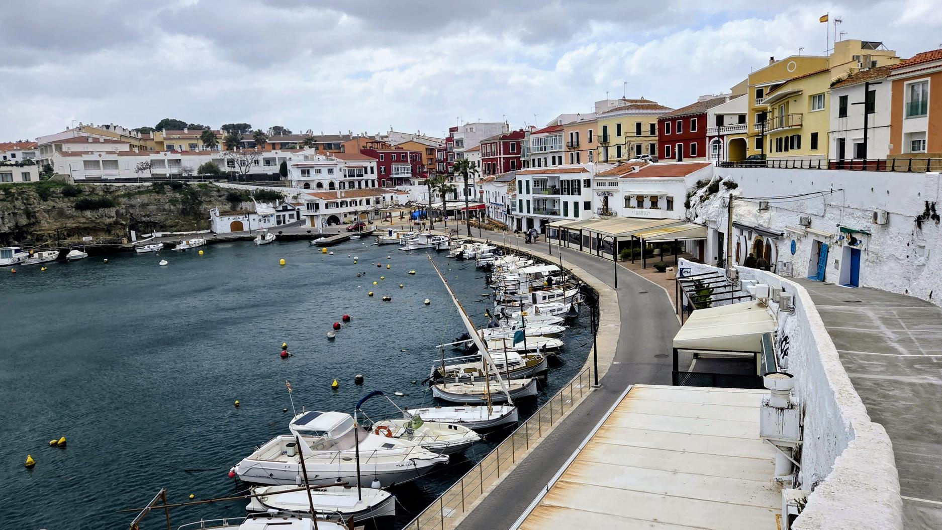A small harbour in Es Castell, Menorca, with calm water, moored boats, and rows of colourful white, red, and yellow buildings lining the waterfront under an overcast sky.